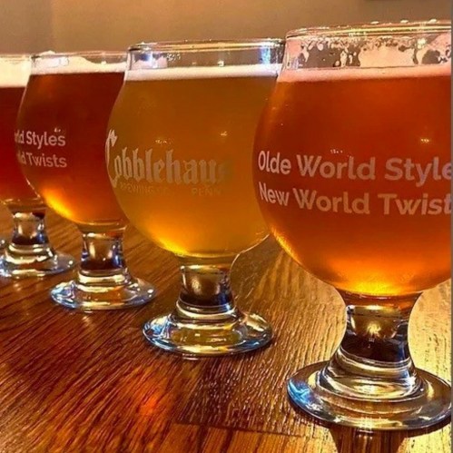 Four glasses of craft beer in varying colors, each labeled with unique names, arranged on a wooden table.