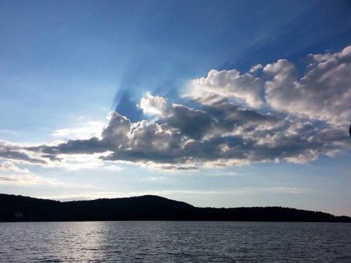 A serene lake scene with a blue sky, fluffy clouds, and sunlight casting rays over distant hills.