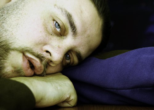A close-up of a man resting his head on his arm, with a thoughtful expression and striking blue eyes.