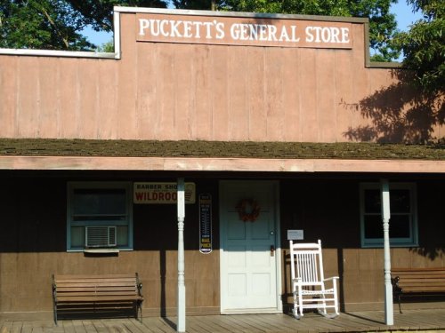A rustic general store with a sign reading "Puckett's General Store," featuring a porch and rocking chair.