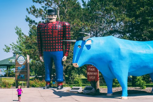 A giant Paul Bunyan statue stands next to a blue ox, with a child walking nearby in a park setting.