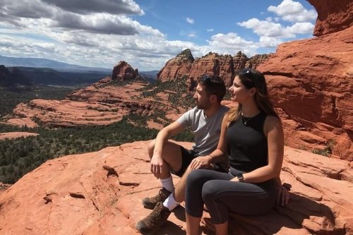 A man and woman sit on a red rock ledge, overlooking a scenic landscape of mountains and trees under a cloudy sky.