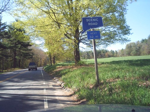 A scenic road sign indicating the next scenic area is 32 miles ahead, surrounded by green trees and grass.