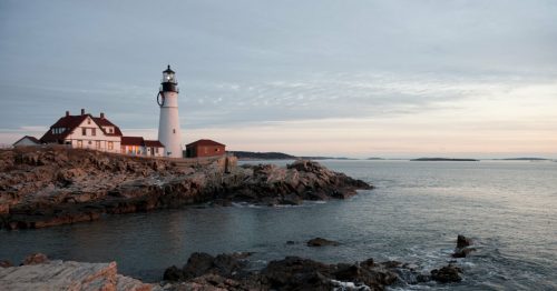 A lighthouse stands on rocky shores at sunset, with a calm sea and distant islands in the background.