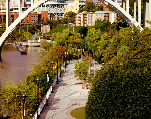 A scenic riverside walkway lined with trees and lampposts, with a bridge arching overhead and buildings in the background.