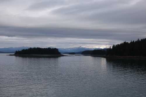 A serene landscape featuring calm waters, distant mountains, and a small island under a cloudy sky.