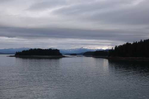 A serene landscape featuring calm waters, a small island, and distant mountains under a cloudy sky.