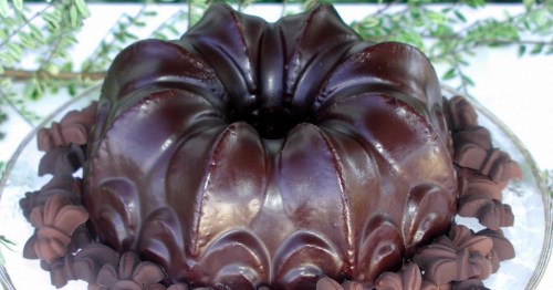 A glossy chocolate bundt cake surrounded by decorative chocolate flowers on a glass platter.