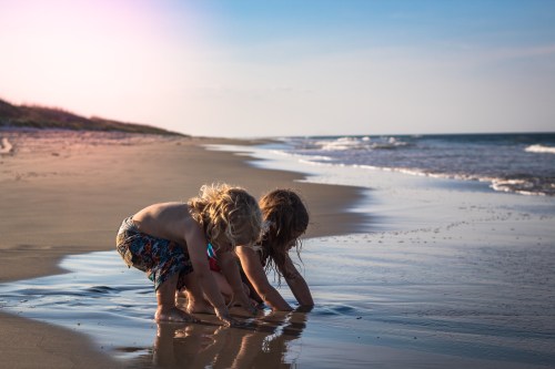 Two children play in shallow water on a beach, enjoying a sunny day with gentle waves in the background.