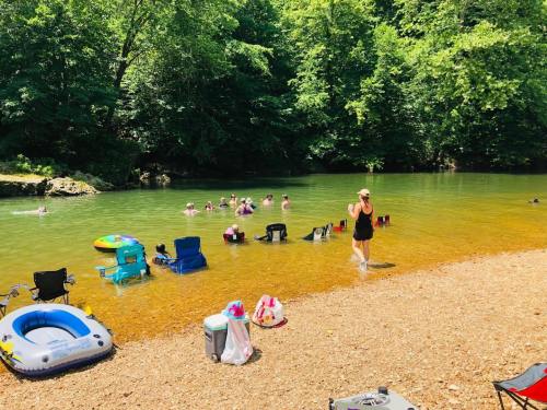 A sunny river scene with people swimming and relaxing on the shore, surrounded by trees and beach chairs.