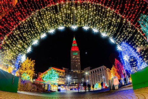 Colorful holiday lights illuminate a city square at night, featuring a tall building with a red and green top.