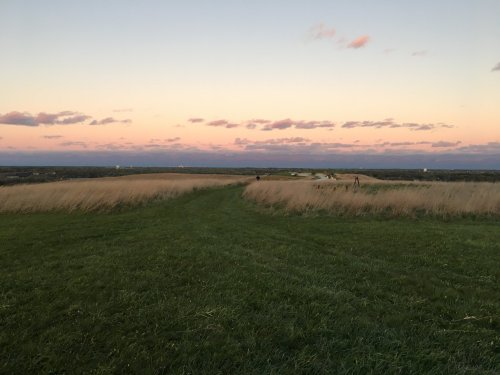 A grassy path winds through tall grass, leading to a colorful sunset sky with scattered clouds.