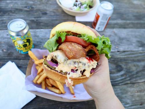 A hand holding a large burger with lettuce, tomato, and onion, alongside fries and drinks on a wooden table.