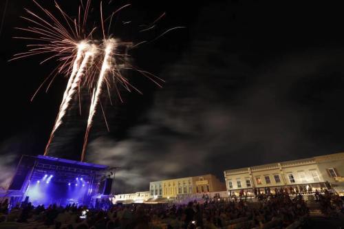 Fireworks illuminate the night sky above a crowd enjoying a concert, with a stage and historic buildings in the background.