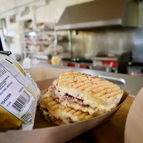 A close-up of a grilled sandwich with layers of meat and cheese, served in a takeout box next to a bag of chips.