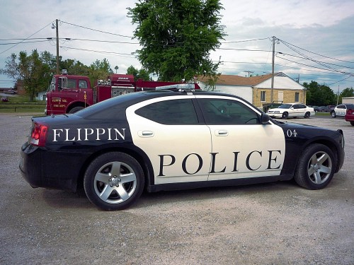 A black and white police car marked "Flippin Police" parked in a gravel lot with trees and buildings in the background.