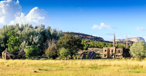 A scenic view of a deserted landscape featuring dilapidated buildings, trees, and rolling hills under a blue sky.