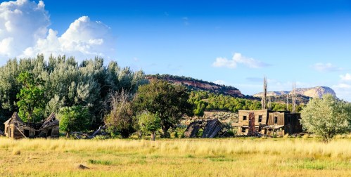 Abandoned buildings amidst tall grass and trees, set against a backdrop of blue sky and distant hills.