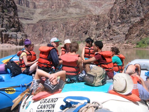 A group of people in life jackets sitting on a raft in the Grand Canyon, surrounded by rocky cliffs and water.