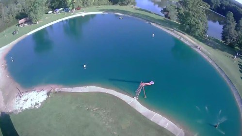 Aerial view of a clear blue pond with a diving platform, surrounded by green grass and trees.