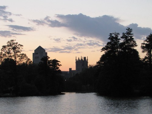 Silhouette of buildings and trees against a sunset sky reflected in a calm lake.