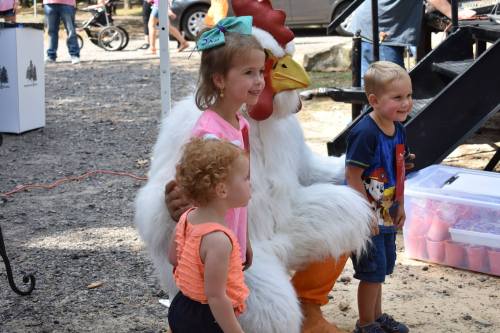Two children pose with a person in a chicken costume at an outdoor event, smiling and enjoying the moment.