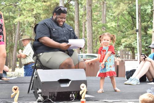 A man sits on a chair reading, while a smiling young girl in a colorful dress dances beside him at an outdoor event.