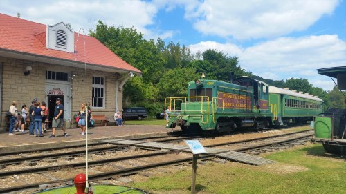A green vintage train at a station with people waiting outside, surrounded by trees and a blue sky.