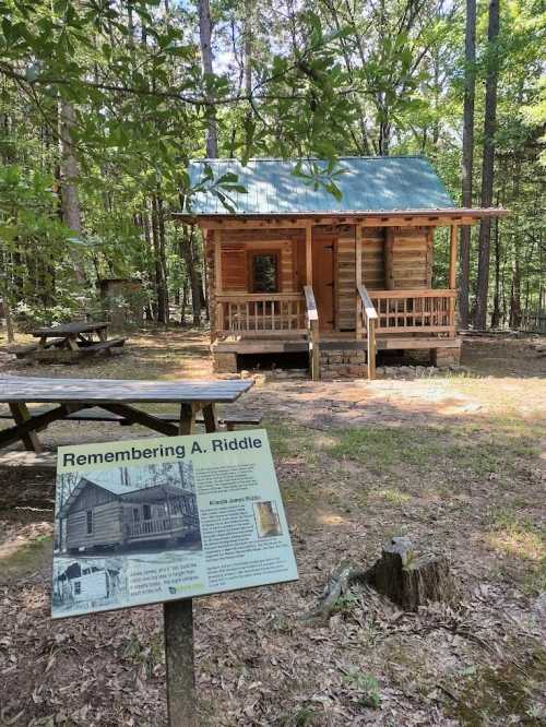 A wooden cabin in a forested area, with a sign about A. Riddle in the foreground and picnic tables nearby.