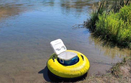 A yellow inflatable tube with a cooler sits on the edge of a calm river, surrounded by grass and water.
