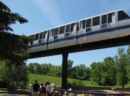 A monorail glides above a park, with people walking below on a sunny day surrounded by greenery.
