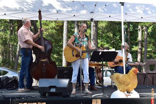 A woman sings and plays guitar on stage, accompanied by musicians with a bass and guitar, with a chicken nearby.