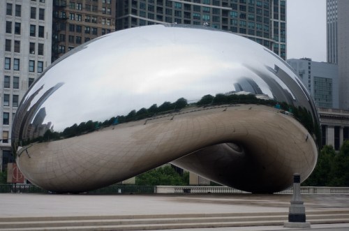 A large, reflective sculpture resembling a bean, set against a city skyline with overcast skies.