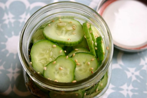 A jar filled with sliced cucumbers and sesame seeds, sitting on a patterned surface with a lid nearby.
