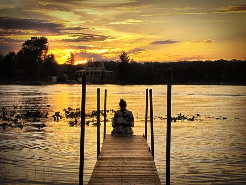 A person sits on a dock by a lake, watching a vibrant sunset with clouds reflecting on the water.
