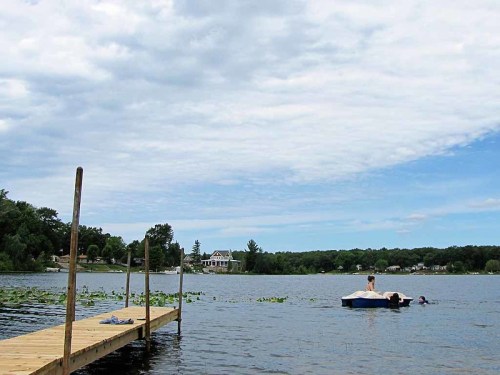 A serene lake scene with a wooden dock, a person on a float, and a house in the background under a cloudy sky.