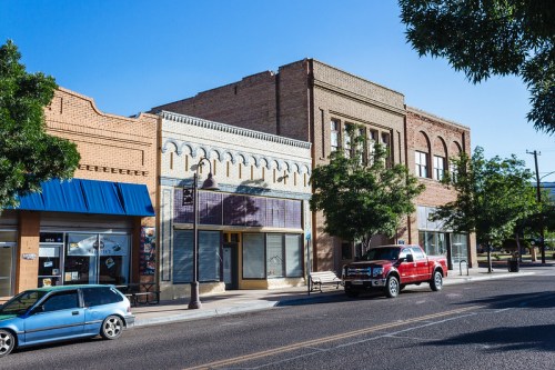 A sunny street scene featuring brick buildings, a blue awning, and parked vehicles along a quiet downtown area.