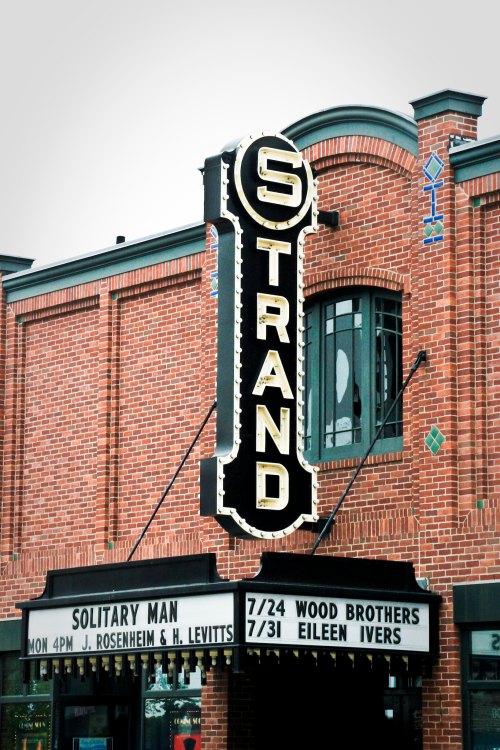 Sign for the Strand theater featuring upcoming events and a marquee for "Solitary Man." Brick building backdrop.