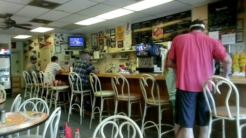 A casual diner interior with patrons at the counter, a TV on the wall, and tables with chairs in the foreground.