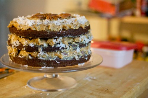 A three-layer chocolate cake topped with coconut and pecans, displayed on a glass cake stand.