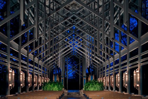 A modern, open chapel with a geometric wooden structure, illuminated by soft lights, surrounded by trees at dusk.