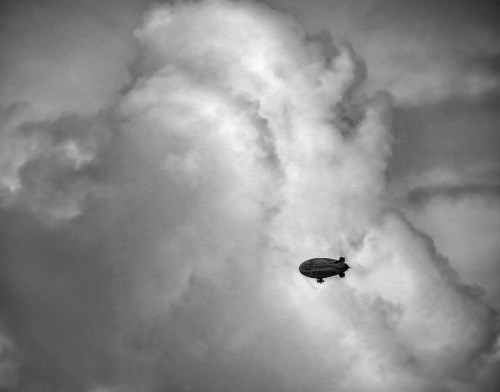 A black and white image of a zeppelin floating among fluffy clouds in a dramatic sky.
