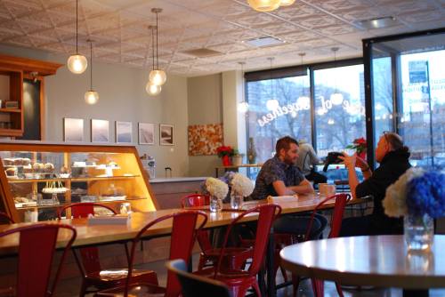 Two people chat at a café with a display case of pastries, surrounded by bright decor and flowers on the tables.