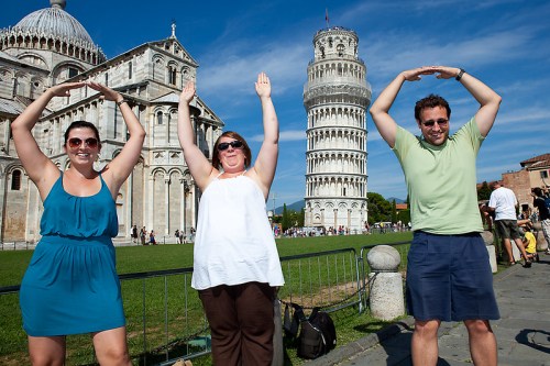 Three people pose with arms raised in front of the Leaning Tower of Pisa, smiling against a clear blue sky.