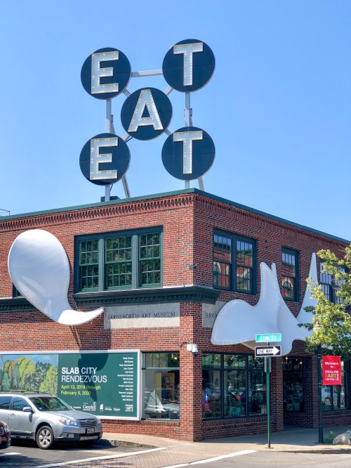 A brick building with a large "EAT" sign on top and whimsical white shapes on the facade, featuring a museum advertisement.