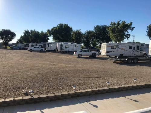 A row of parked RVs and a car on a gravel lot under a clear blue sky, surrounded by trees.