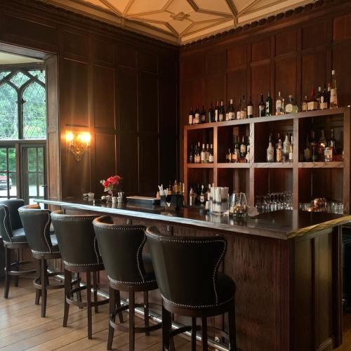 A cozy bar with dark wood paneling, a well-stocked shelf of bottles, and elegant seating.