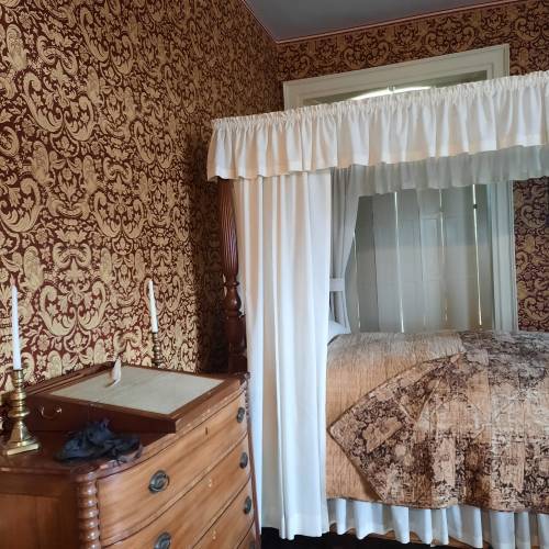 A vintage bedroom featuring a four-poster bed with white drapes, ornate wallpaper, and a wooden dresser.