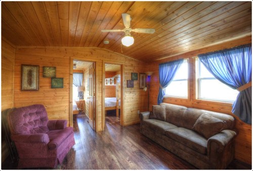 Cozy wooden interior of a cabin with a sofa, armchair, and windows draped with blue curtains.