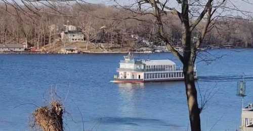 A riverboat cruises on a calm lake, surrounded by trees and distant houses on the shore.